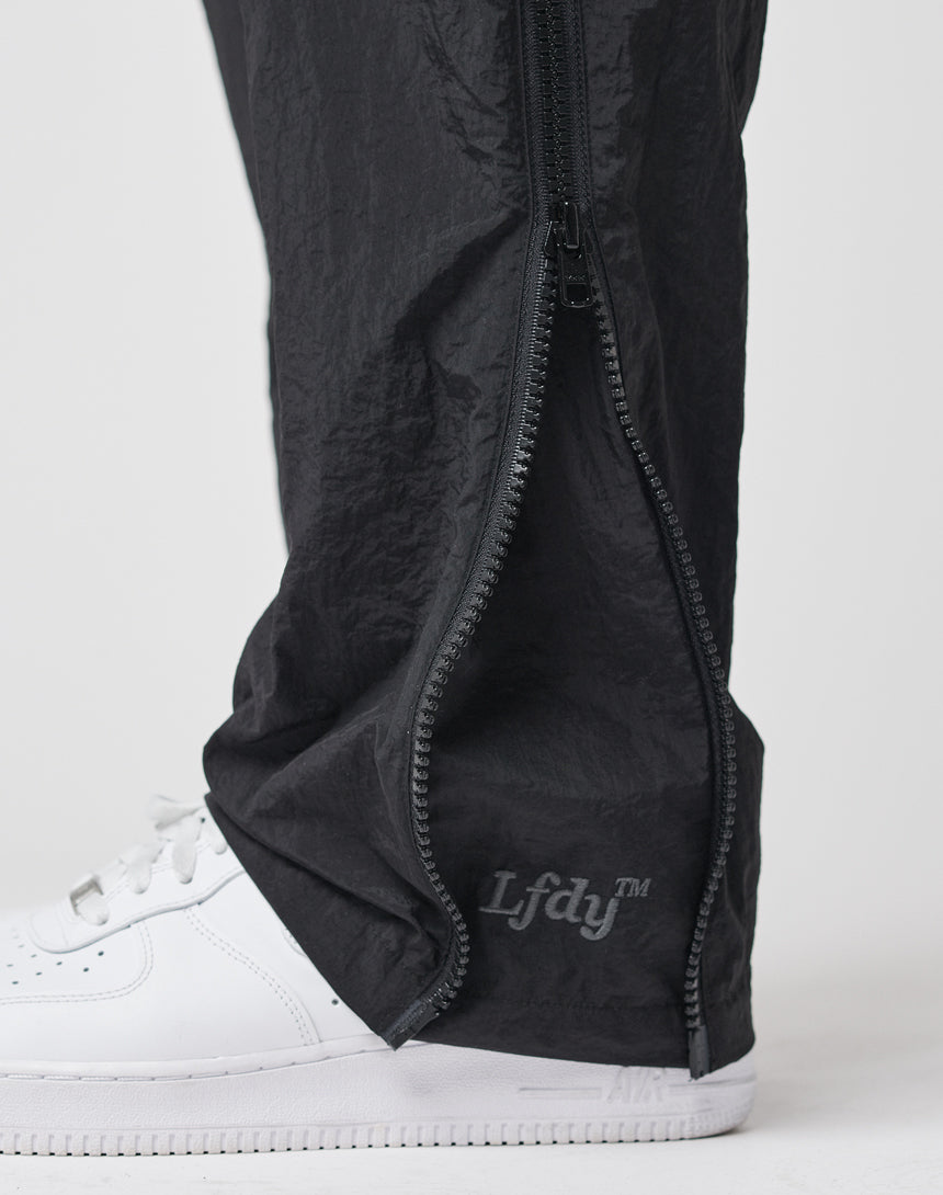 Close-up of a person wearing LFDY Zip Tape Track Pants in black nylon, with the ankle zipper partially open to show a white sneaker and the LFDY logo near the ankle.