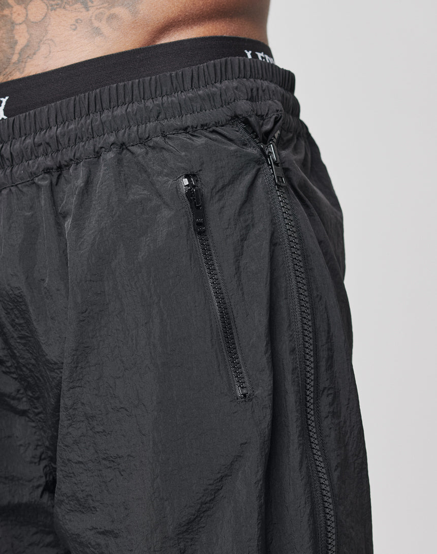 Close-up of a persons waist wearing LFDY Zip Tape Track Pants in black nylon with a crinkled texture, elastic waistband, and visible zippers on the fly and side pocket; part of their tattooed torso is also visible.