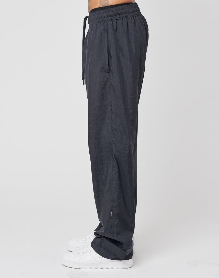 A person in profile wears LIVE FAST DIE YOUNG Nylon Jogger—regular fit, black nylon pants with a wide elastic waistband and drawstring—paired with white sneakers; a tattoo is visible on their lower torso against a plain white background.