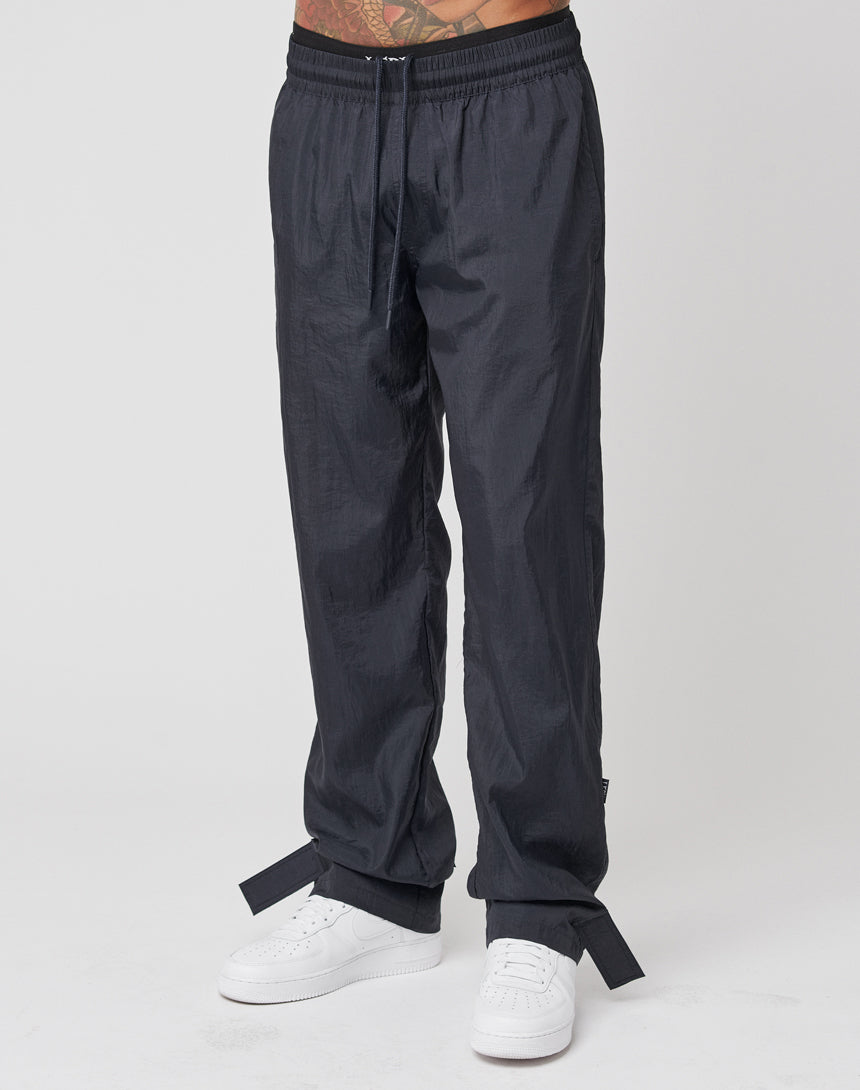 A person is shown from the waist down wearing LIVE FAST DIE YOUNG Nylon Jogger pants in black with a wide elastic waistband, drawstring, ankle straps, and white sneakers against a plain white background.