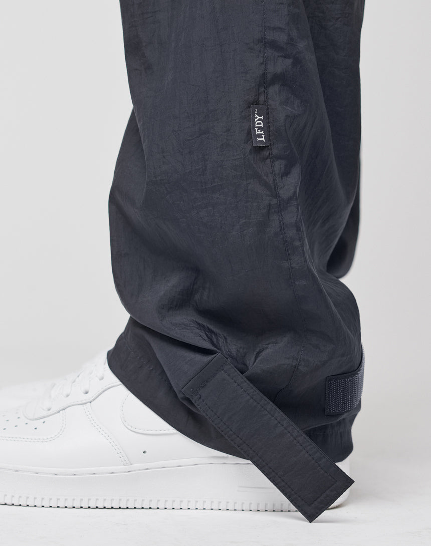 A close-up of a person wearing LIVE FAST DIE YOUNG Nylon Jogger pants in black nylon with a regular fit, JFPV tag, ankle velcro strap, and crisp white sneakers, set against a plain background.