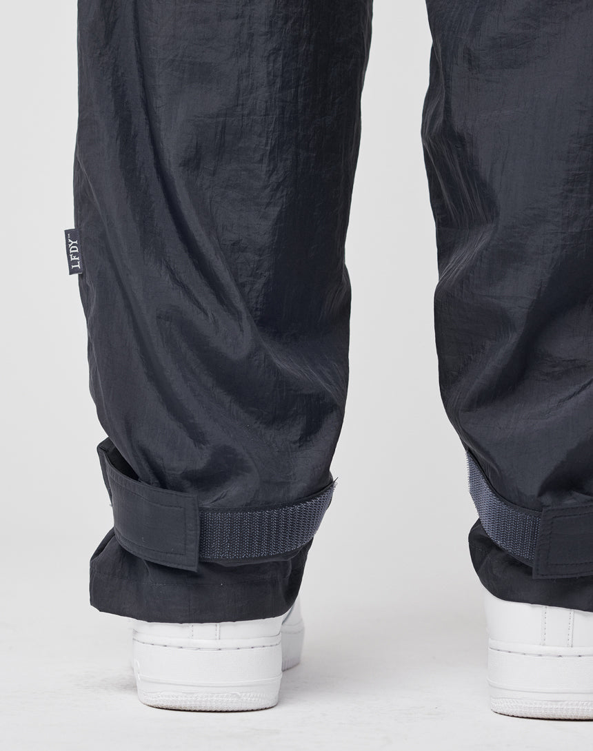 Close-up of a person wearing LIVE FAST DIE YOUNG Nylon Jogger—regular fit black nylon pants with an elastic waistband and adjustable cuff straps—paired with white sneakers, against a plain white background.