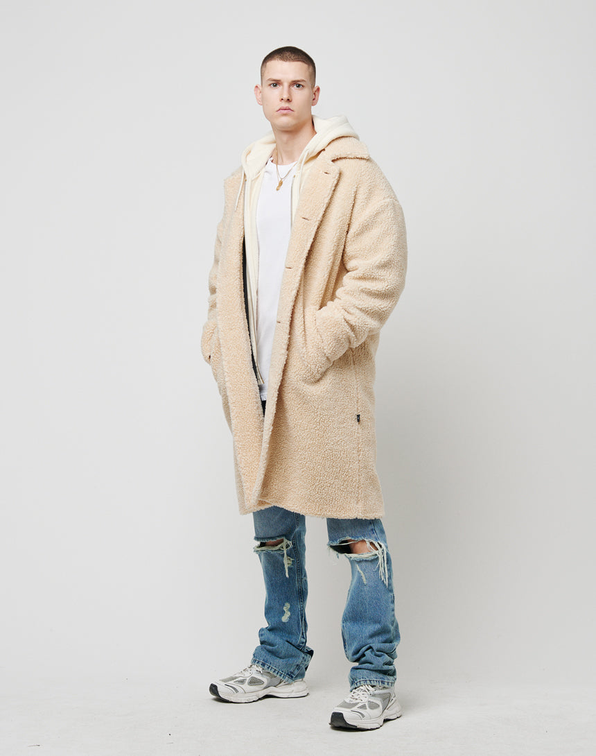 A young man stands against a white background, wearing the LFDY Sherpa Coat by LFDY in one size over a white hoodie and T-shirt, distressed blue jeans with knee rips, and gray sneakers. His hands are in the coat pockets.