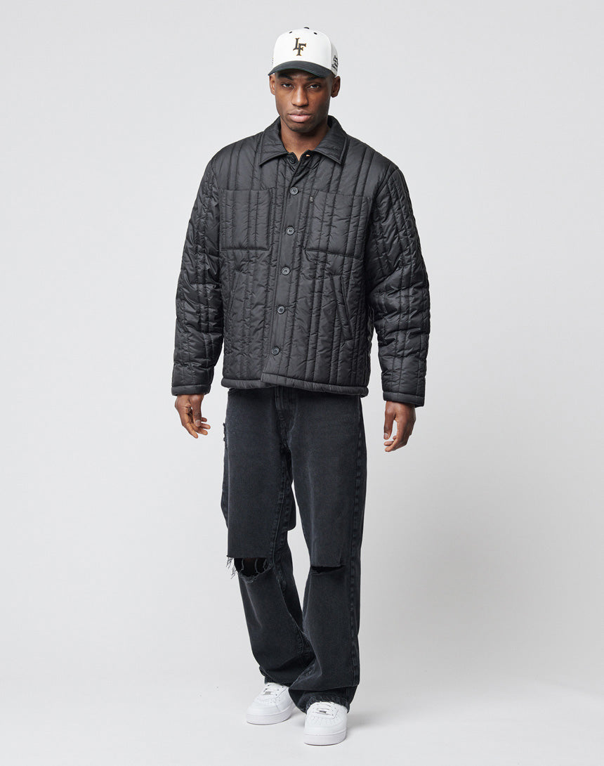 A man wears the Import & Export Jacket by LIVE FAST DIE YOUNG, paired with black distressed jeans and white sneakers, standing against a plain light gray background.