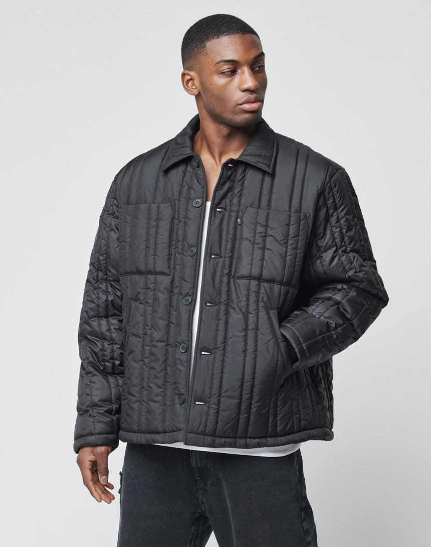 A man wears a black Import & Export Jacket by LIVE FAST DIE YOUNG over a white shirt, standing against a plain light gray background. He gazes slightly left, with one hand by his side and the other partially in his pocket.