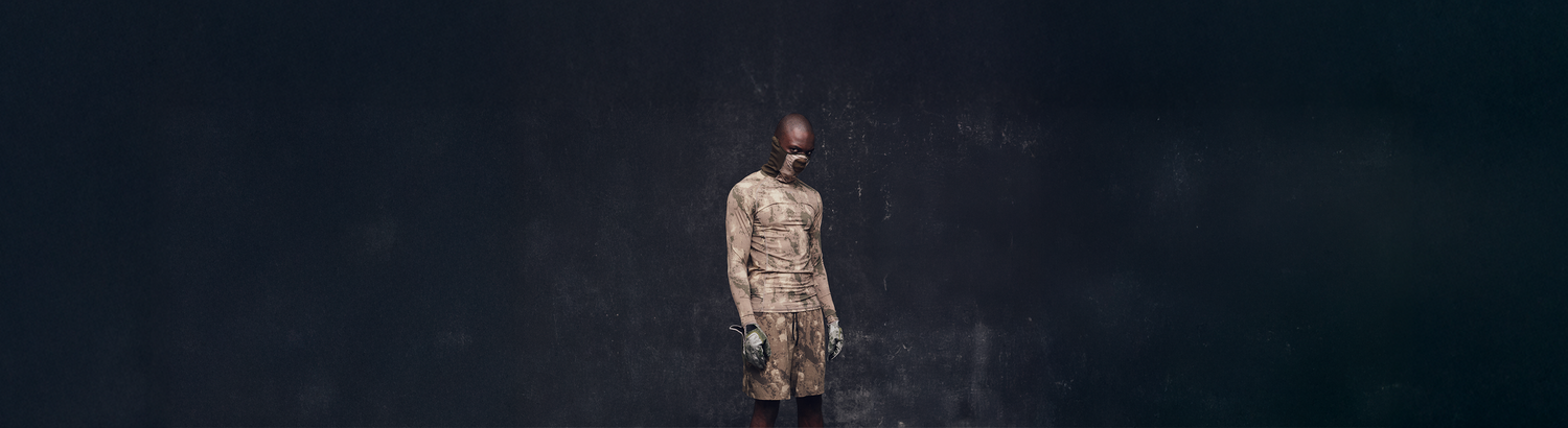 A person wearing a matching earth-toned mask, long-sleeve shirt, and shorts stands against a dark, textured background, looking downward with their arms at their sides.