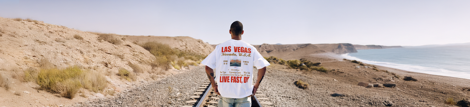A person stands on railroad tracks facing away, wearing a white Las Vegas T-shirt, surrounded by desert landscape with hills and a coastline under a clear sky.