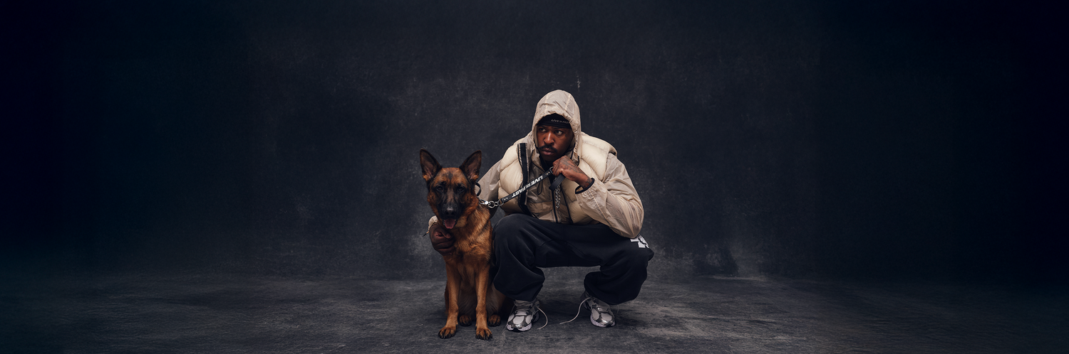 A man wearing a light-colored hooded jacket crouches next to a German Shepherd dog on a leash, both looking toward the camera against a dark background.