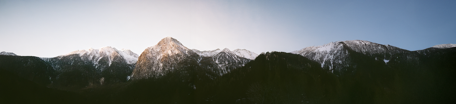 Panoramic view of snow-capped mountain peaks under a clear blue sky, with sunlight illuminating the tallest peak and the lower slopes in shadow.