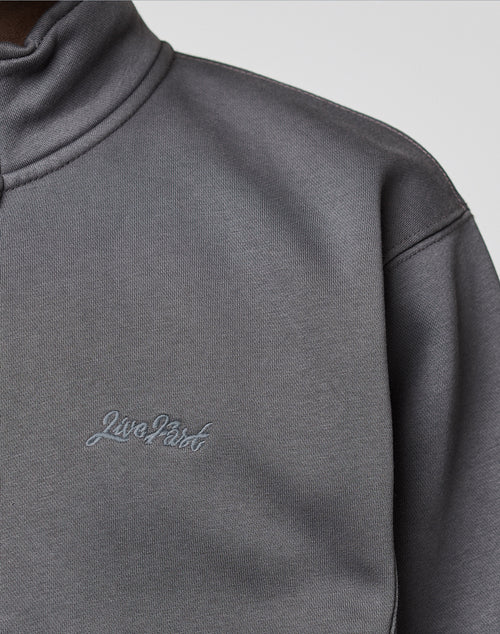 Close-up of a person wearing a grey LIVE FAST DIE YOUNG Sweatsuit Jacket with a standing collar and "Live First" embroidered on the chest in grey thread.