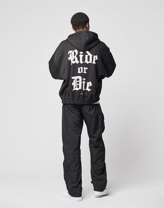 A person faces away, wearing the LIVE FAST DIE YOUNG Ride or Die Hooded Zip with doppellagige Kapuze and bold white “Ride or Die” gothic letters, paired with loose black pants (elastischer Bund) and white sneakers, against a white background.