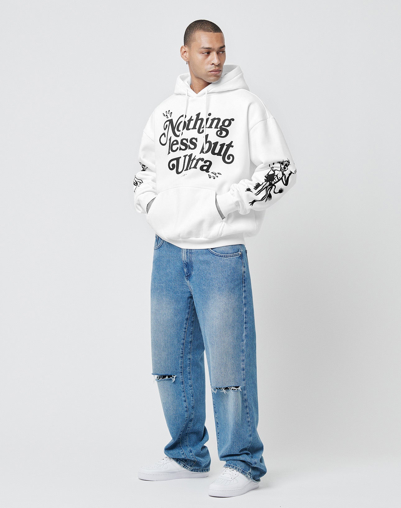 A person in a white LIVE FAST DIE YOUNG LFDY Ultra Hooded with "Nothing less but Ultra" on the front, Bündchen an Ärmeln, and oversize fit, styled with light blue ripped jeans and white sneakers, stands against a plain background looking sideways.