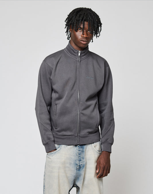 A young man with short dreadlocks wears the LIVE FAST DIE YOUNG Sweatsuit Jacket in dark gray with Stehkragen and Regular Fit, paired with light blue jeans, standing against a plain light gray background looking neutral.