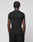 A person with short, twisted hair is shown from behind in a plain black LIVE FAST DIE YOUNG LFDY Slim Tee and black pants against a light gray background. A small Flag-Label detail completes the minimalist look.