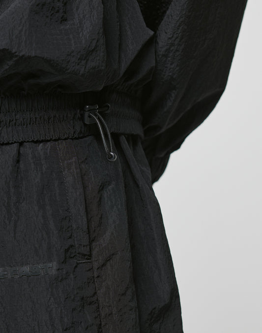 Close-up of a person wearing the LIVE FAST DIE YOUNG Live Fast Nylon Halfzip—a black, textured, crinkled polyester jacket and pants with an elastic waistband and drawstring in an oversized fit—set against a plain light background.