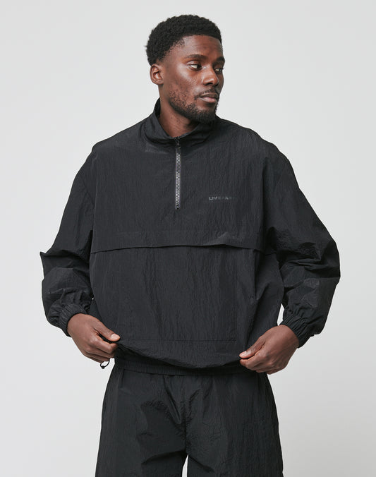 A man stands against a plain background wearing the LIVE FAST DIE YOUNG Live Fast Nylon Halfzip, an oversized black zip-up pullover jacket, paired with matching black pants. He looks slightly left, holding the sides of his jacket.