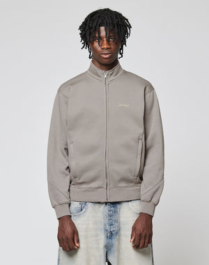 A young man with dark curly hair wears a LIVE FAST DIE YOUNG Sweatsuit Jacket in light gray and faded blue jeans, standing against a plain light gray background with a neutral expression.