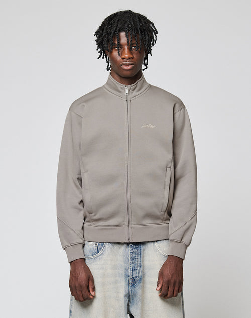 A young man with dark curly hair wears a LIVE FAST DIE YOUNG Sweatsuit Jacket in light gray and faded blue jeans, standing against a plain light gray background with a neutral expression.