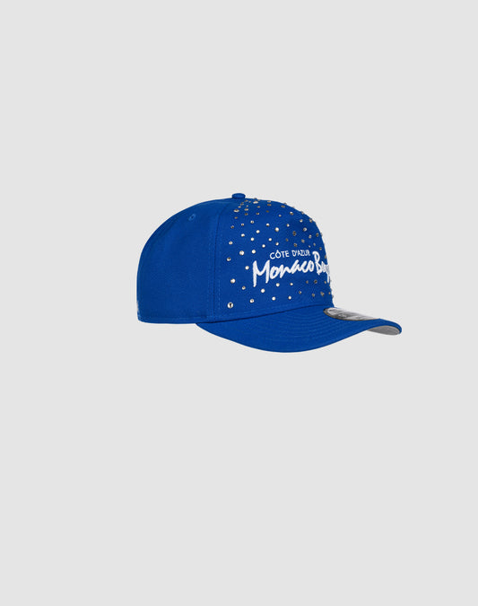 The Custom LFDY X NEW ERA Monaco Boys cap by LIVE FAST DIE YOUNG features silver studs and white Côte D’Azur Monaco Blue embroidery on blue, handdecorated cap style, displayed against a plain light gray background.