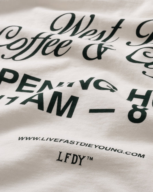 A close-up of the Live Fast Coffee Tee by LIVE FAST DIE YOUNG shows its slightly wrinkled white cotton fabric with black print: West Coffee & LFDY Berlin Café, hours 7AM–8PM, website address, “LFDY™,” and made from 100% Baumwolle.