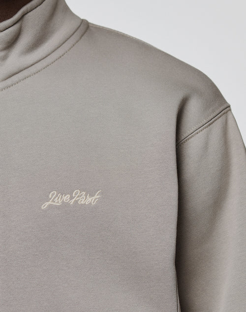 A close-up of a person wearing a light grey LIVE FAST DIE YOUNG Sweatsuit Jacket, Regular Fit, with "Live Fast" embroidered in small white cursive letters on the chest.