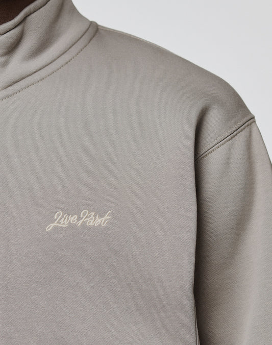 A close-up of a person wearing a light grey LIVE FAST DIE YOUNG Sweatsuit Jacket, Regular Fit, with "Live Fast" embroidered in small white cursive letters on the chest.