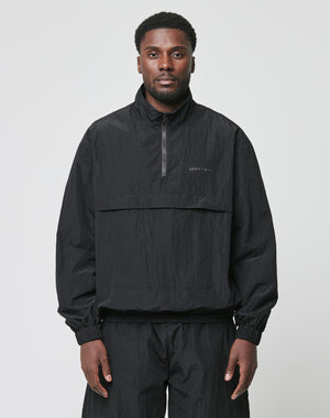 A man faces forward against a light gray background, wearing the LIVE FAST DIE YOUNG Live Fast Nylon Halfzip—an oversized black, long-sleeve, half-zip pullover jacket—paired with matching black polyester pants.