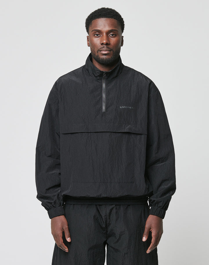 A man faces forward against a light gray background, wearing the LIVE FAST DIE YOUNG Live Fast Nylon Halfzip—an oversized black, long-sleeve, half-zip pullover jacket—paired with matching black polyester pants.