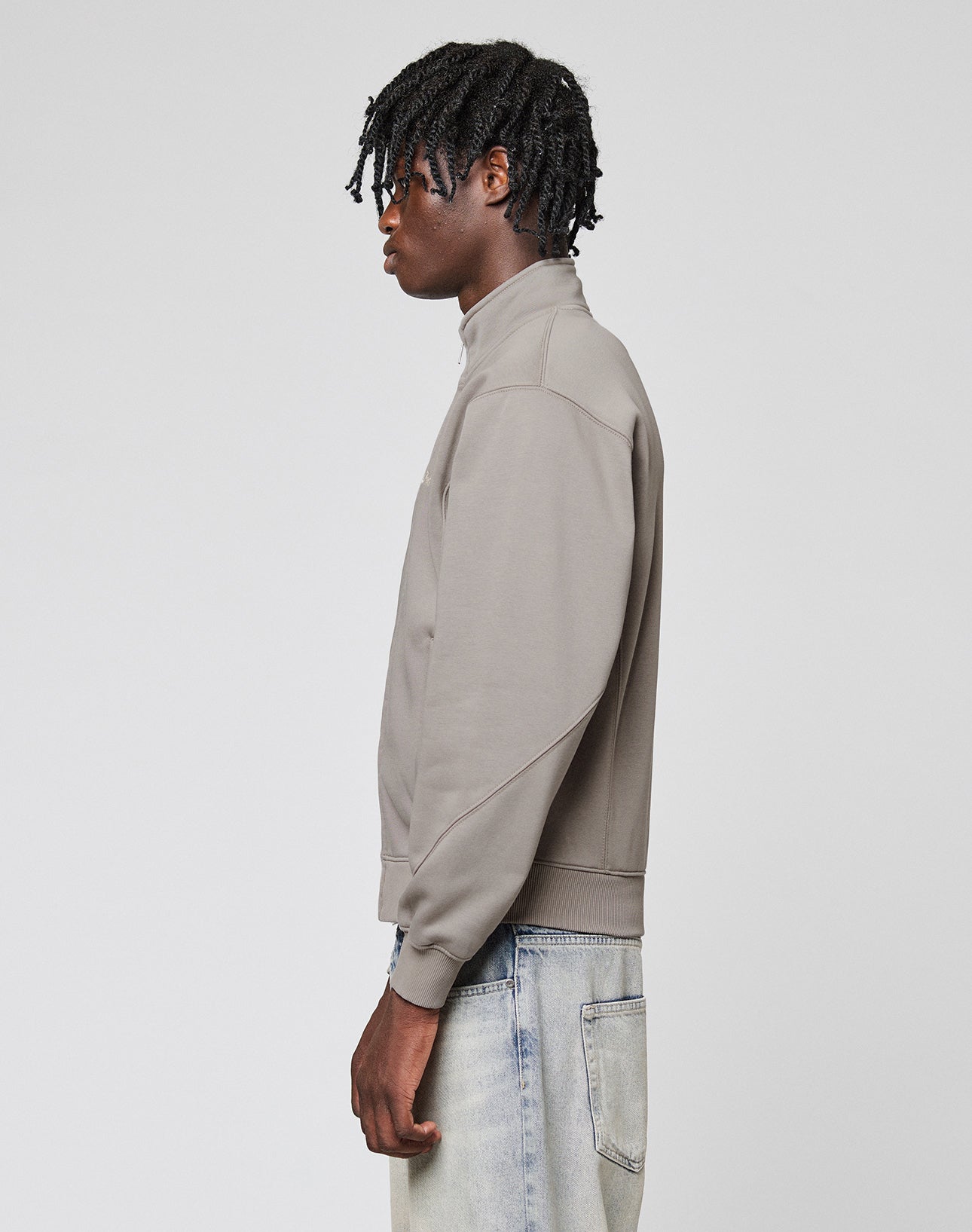 A young man with short, twisted hair stands in profile against a plain background, wearing the LIVE FAST DIE YOUNG Sweatsuit Jacket in taupe with Stehkragen and Regular Fit light blue jeans.