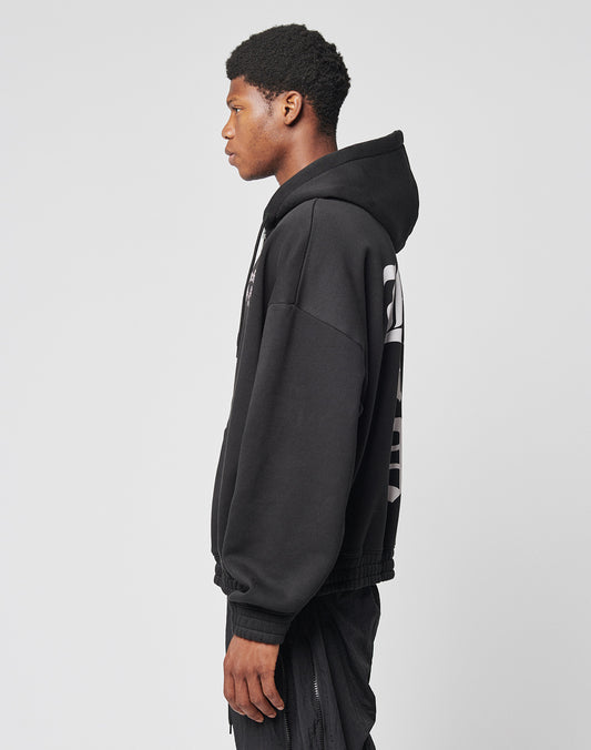 A young man stands in profile against a plain background, wearing the LIVE FAST DIE YOUNG Ride or Die Hooded Zip in black with an oversize fit, doppellagige Kapuze, black pants, and a bold white back design.