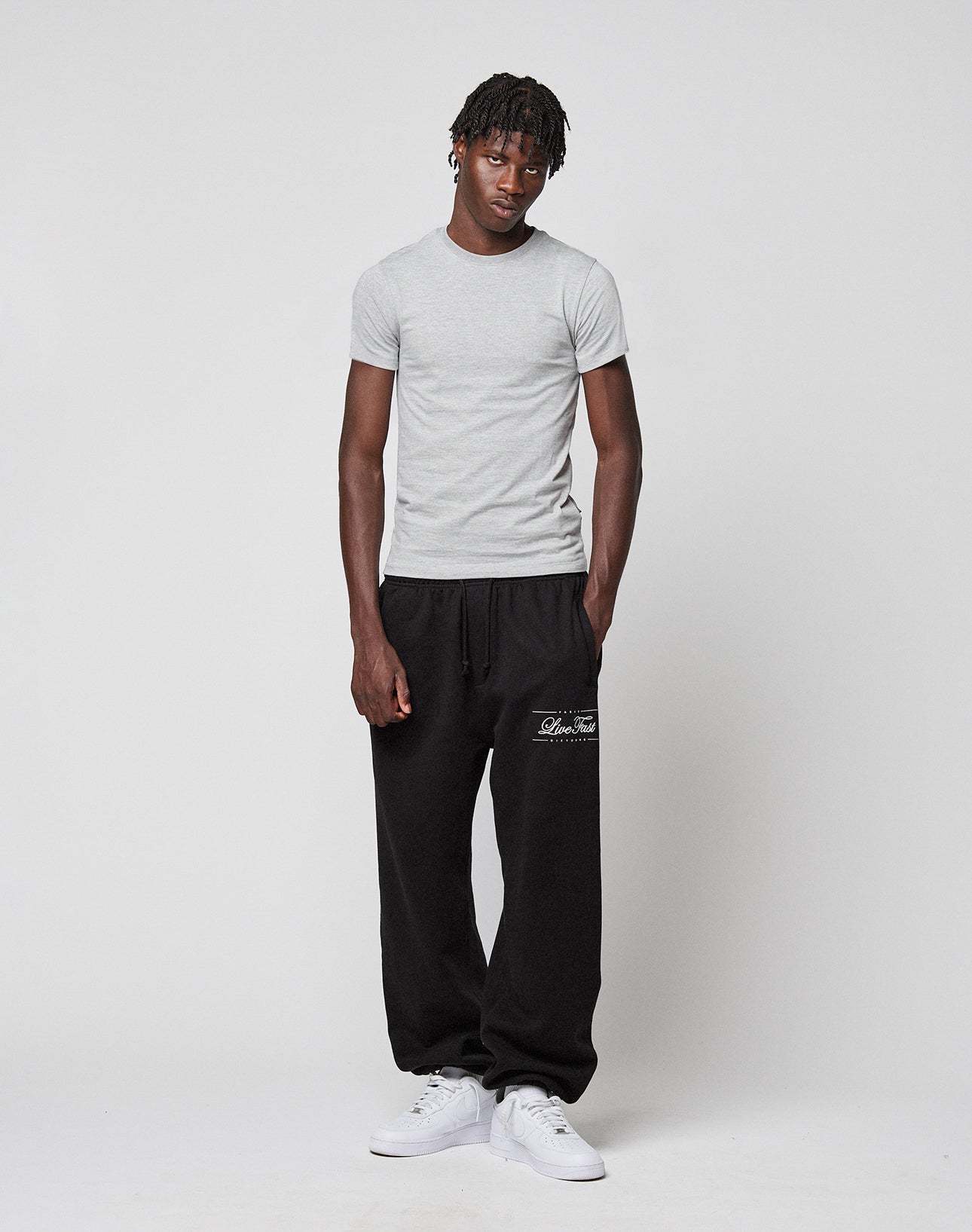 A man wears the LIVE FAST DIE YOUNG LFDY Slim Tee in light gray cotton, loose black sweatpants with white text on the left leg, and white sneakers. His left hand is in his pocket as he faces the camera. A Flag-Label detail completes the look.