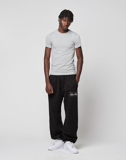 A man wears the LIVE FAST DIE YOUNG LFDY Slim Tee in light gray cotton, loose black sweatpants with white text on the left leg, and white sneakers. His left hand is in his pocket as he faces the camera. A Flag-Label detail completes the look.