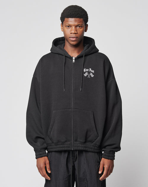 A young man wearing the LIVE FAST DIE YOUNG Ride or Die Hooded Zip—a black hoodie with doppellagige Kapuze, front pockets, and white chest text and graphic—stands facing forward against a plain light gray background.
