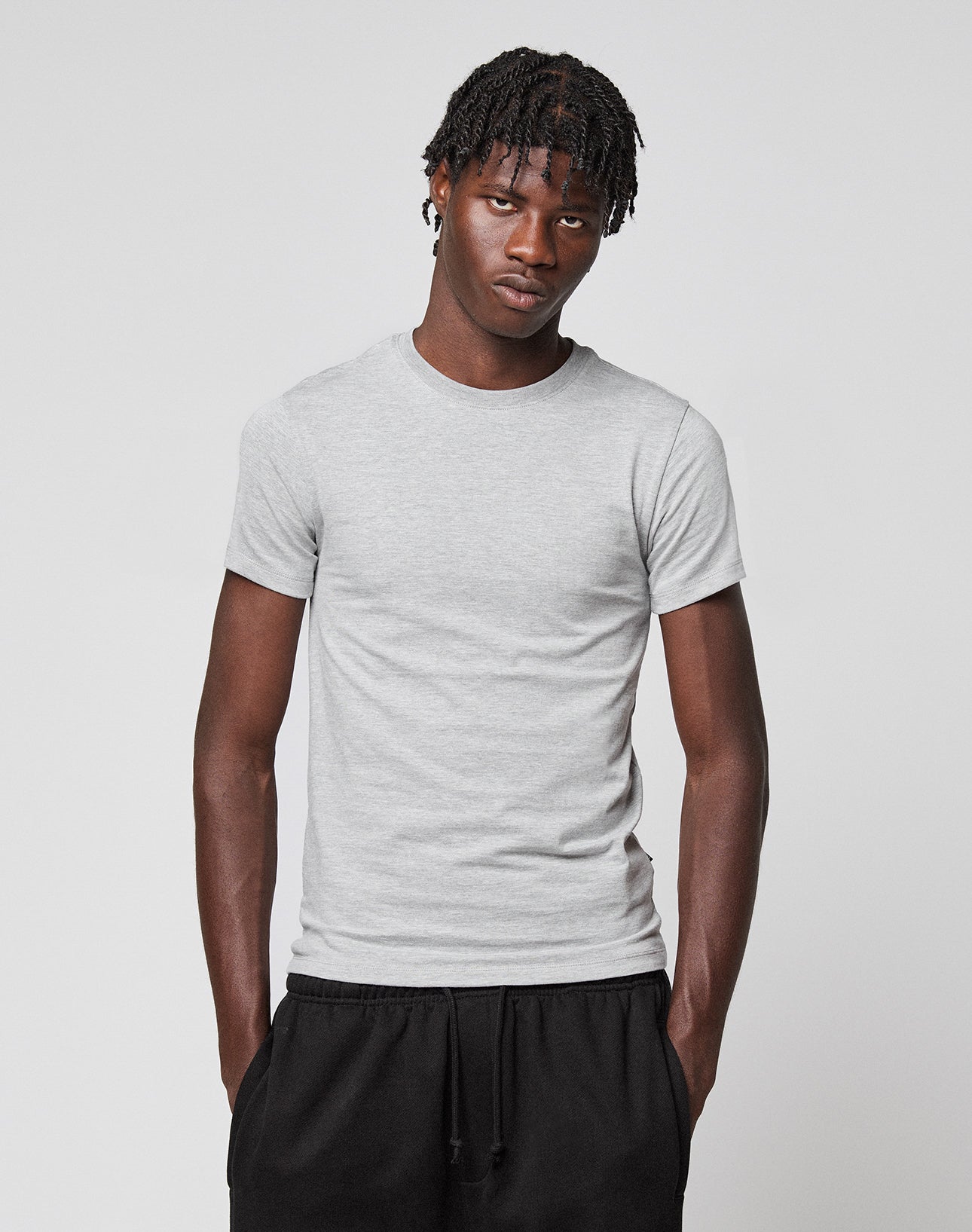 A young man with short dreadlocks wears the LIVE FAST DIE YOUNG LFDY Slim Tee in light gray and black pants, standing against a plain light background with his hands in his pockets, looking slightly downward.