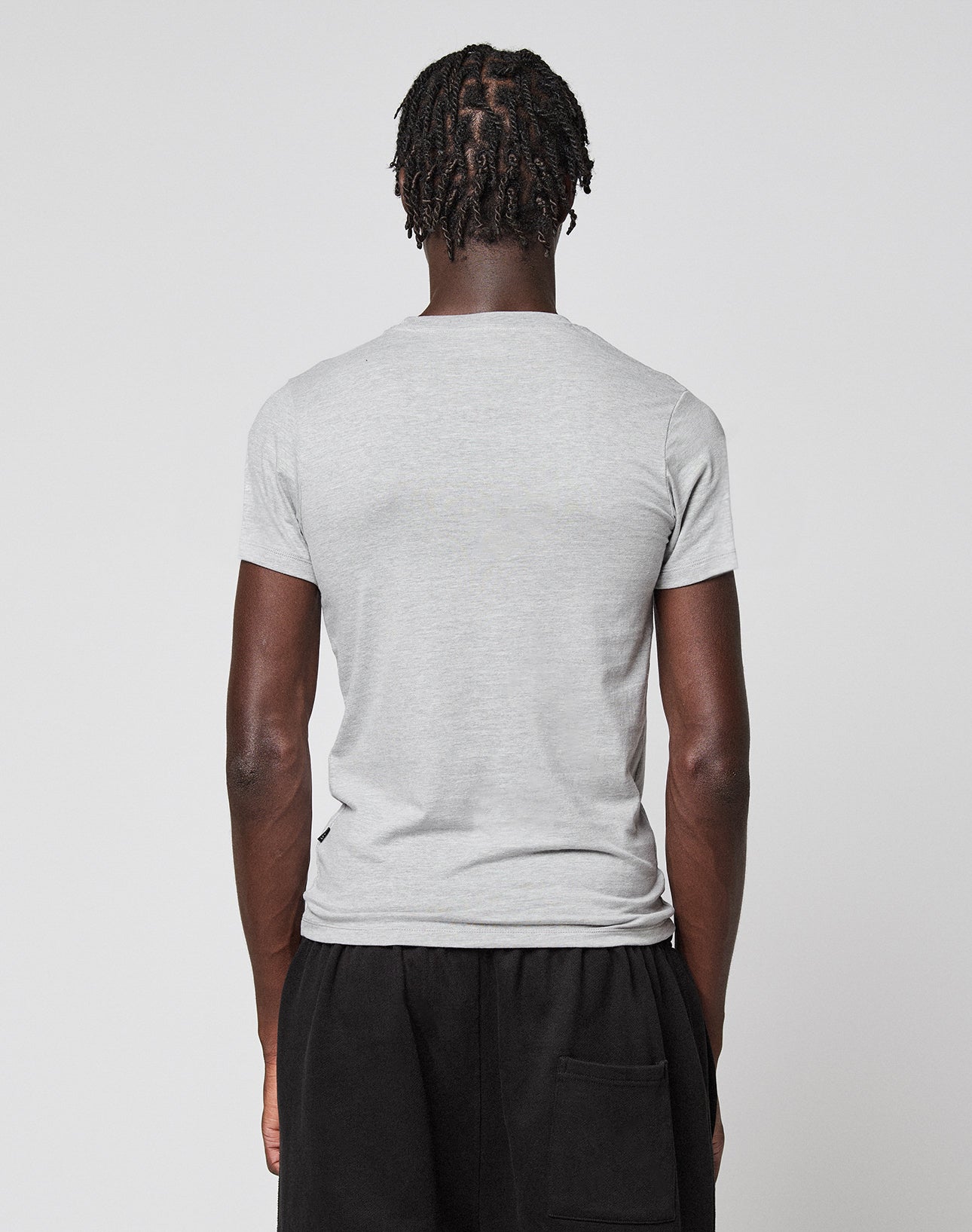 A person with short, twisted hair stands with their back to the camera, wearing a light gray LFDY Slim Tee by LIVE FAST DIE YOUNG and black pants. The tee features a subtle Flag-Label and is set against a plain light background.