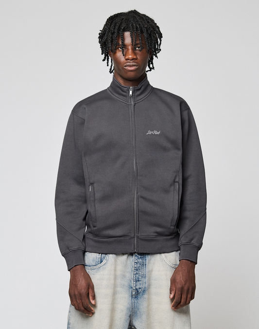 A young man with short dreadlocks wears a LIVE FAST DIE YOUNG Sweatsuit Jacket in dark gray and light blue jeans, standing against a plain light gray background and looking directly at the camera with a neutral expression. | model