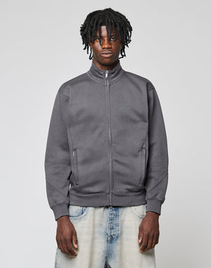 A young man with short dreadlocks wears a LIVE FAST DIE YOUNG Sweatsuit Jacket in dark gray and light blue jeans, standing against a plain light gray background and looking directly at the camera with a neutral expression.