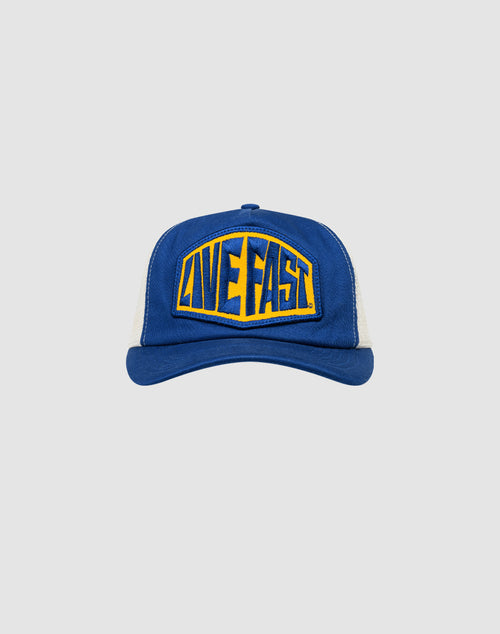 The LFDY Patched Trucker by LIVE FAST DIE YOUNG is a blue and white cap with a yellow and blue LIVE FAST patch on the front, bold stylized lettering, and an adjustable Snapverschluss closure.