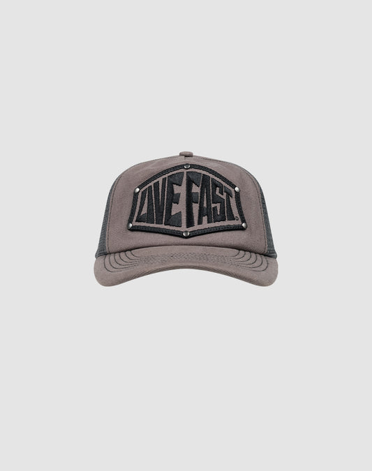 The LFDY Rivet Trucker by LIVE FAST DIE YOUNG is a gray cap with a black mesh back, LOVE FAST patch in bold black letters, silver stud accents, and a subtle flag-label detail.
