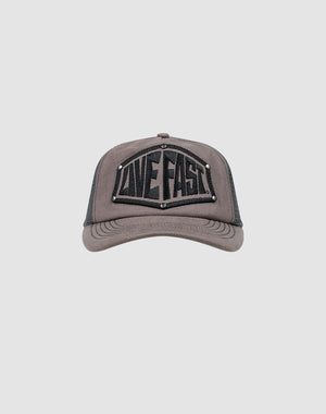 The LFDY Rivet Trucker by LIVE FAST DIE YOUNG is a gray cap with a black mesh back, LOVE FAST patch in bold black letters, silver stud accents, and a subtle flag-label detail.