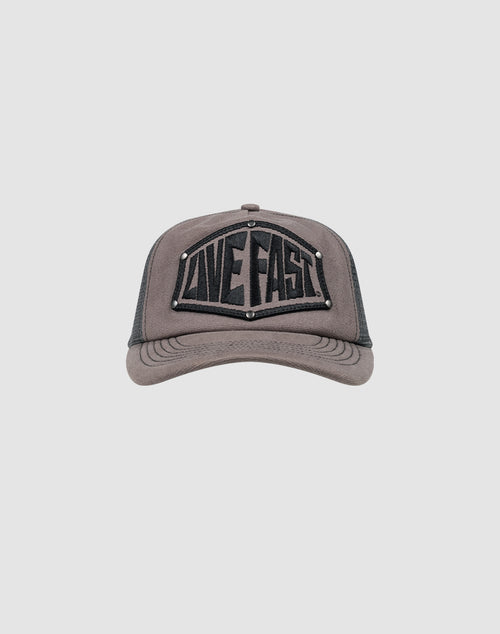 The LFDY Rivet Trucker by LIVE FAST DIE YOUNG is a gray cap with a black mesh back, LOVE FAST patch in bold black letters, silver stud accents, and a subtle flag-label detail.