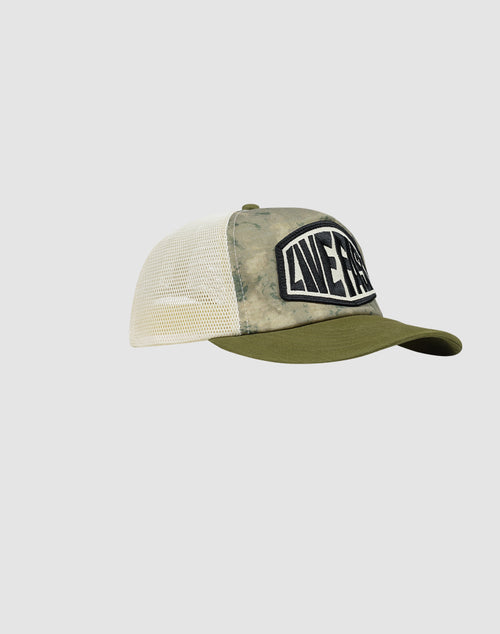 The LFDY Patched Trucker by LIVE FAST DIE YOUNG is a green and beige cap with a mesh back, snap closure, curved brim, and a bold black-and-white LIVEST logo patch on the front. The hat is shown against a plain light background.