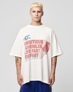 A man with short blond hair wears the LFDY Juveniles Tee by LIVE FAST DIE YOUNG, an oversized 100% Baumwolle shirt with red and blue “AMBITIOUS JUVENILES LIVE FAST DIE FAST” graphics, standing against a plain light background.