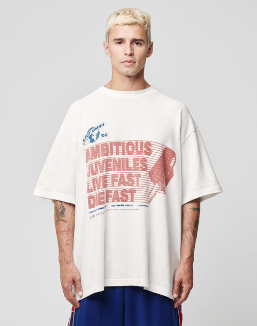 A man with short blond hair wears the LFDY Juveniles Tee by LIVE FAST DIE YOUNG, an oversized 100% Baumwolle shirt with red and blue “AMBITIOUS JUVENILES LIVE FAST DIE FAST” graphics, standing against a plain light background.