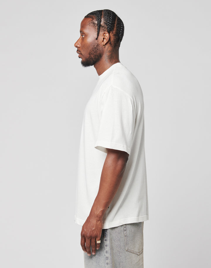 A man with braided hair, wearing a LIVE FAST DIE YOUNG LFDY Vintage Tee in an oversize fit and light gray jeans, stands in profile against a plain light gray background.