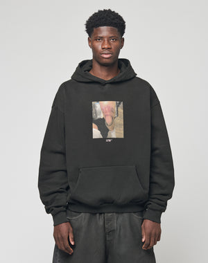 A man with short curly hair wears the LIVE FAST DIE YOUNG LFDY Sharkhunter Hooded in black, featuring a front graphic, standing against a plain light gray background.