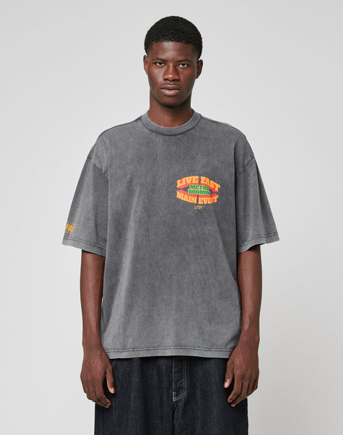A young man with short hair and a neutral expression models the LIVE FAST DIE YOUNG LFDY Metro Boomin Tee, an oversize faded gray 100% cotton shirt with yellow and red sleeve print, paired with wide dark pants against a plain background.