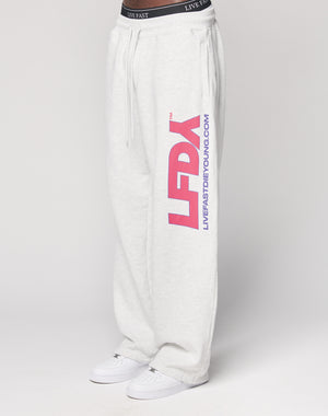 Someone wears the LIVE FAST DIE YOUNG LFDY Racing Wide Jogger in light gray, featuring a wide elastic waistband with LIVE FAST, large pink LFDY letters, and blue LIVEFASTDIEYOUNG.COM on the left leg, styled with white sneakers.