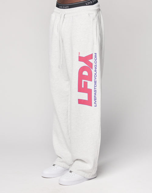Someone wears the LIVE FAST DIE YOUNG LFDY Racing Wide Jogger in light gray, featuring a wide elastic waistband with LIVE FAST, large pink LFDY letters, and blue LIVEFASTDIEYOUNG.COM on the left leg, styled with white sneakers.