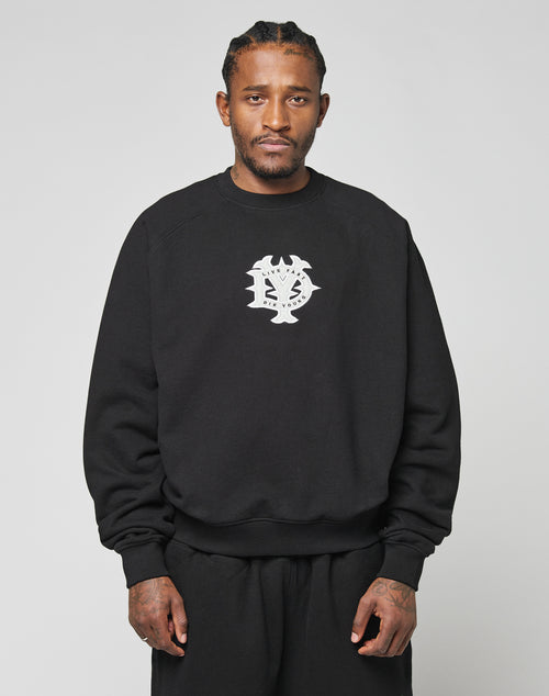 A man with braided hair wears the oversize DY Sweater by LIVE FAST DIE YOUNG, featuring black raglan sleeves and a white chest graphic. He stands against a plain light background with a neutral expression.
